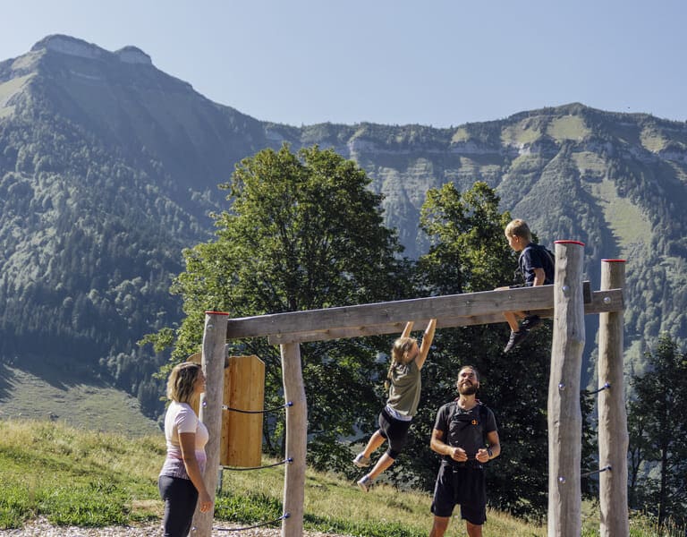 Ein Junge sitz auf dem Klettergerüst, ein Mädchen hangelt sich hinüber, im Hintergrund das Bergpanorama des Schmittenstein