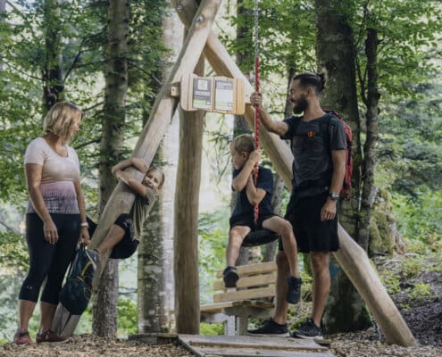 Ein Junge sitz auf der Seilrutsche, der Vater hält das Seil, ein Mädchen klettert auf einen Baumstamm