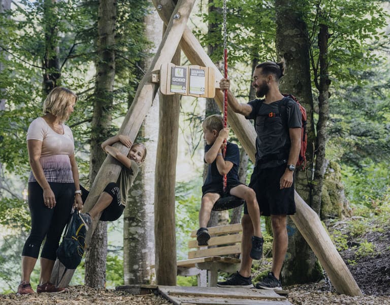 Ein Junge sitz auf der Seilrutsche, der Vater hält das Seil, ein Mädchen klettert auf einen Baumstamm