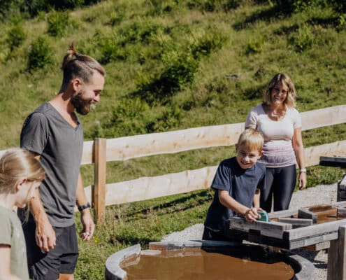 Ein Junge steht an der Wasserspielestation und staut das Wasser, eine Frau, ein Mann und ein Mädchen sehen ihm dabei zu