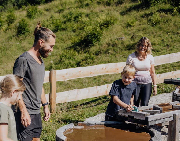 Ein Junge steht an der Wasserspielestation und staut das Wasser, eine Frau, ein Mann und ein Mädchen sehen ihm dabei zu