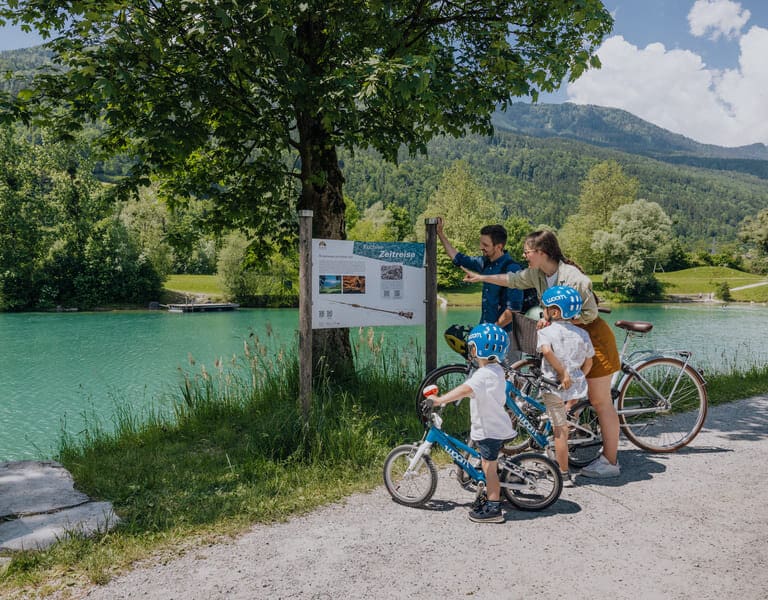 Eine Familie bestehend aus Mann, Frau und 2 kleinen Jungen steht mit den Fahrrädern vor einer Infotafel zum Zeitreiseweg in Kuchl