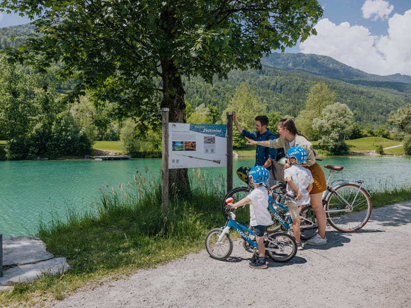 Eine Familie bestehend aus Mann, Frau und 2 kleinen Jungen steht mit den Fahrrädern vor einer Infotafel zum Zeitreiseweg in Kuchl