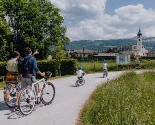 Eine Familie erkundet den Kuchler Zeitreiseweg am Bürgerausee, im Hintergrund ist die Pfarrkirche von Kuchl zu sehen.