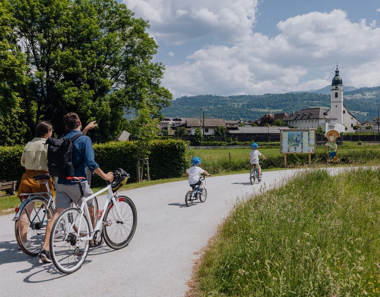 Eine Familie erkundet den Kuchler Zeitreiseweg am Bürgerausee, im Hintergrund ist die Pfarrkirche von Kuchl zu sehen.