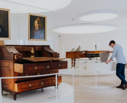 Im Vordergrund ist der Schreibtisch von Franz Xaver Gruber, sowie die Porträts von ihm und seiner Frau zu sehen. Im Hintergrund öffnet ein Besucher eine Schauklappe im Museum.