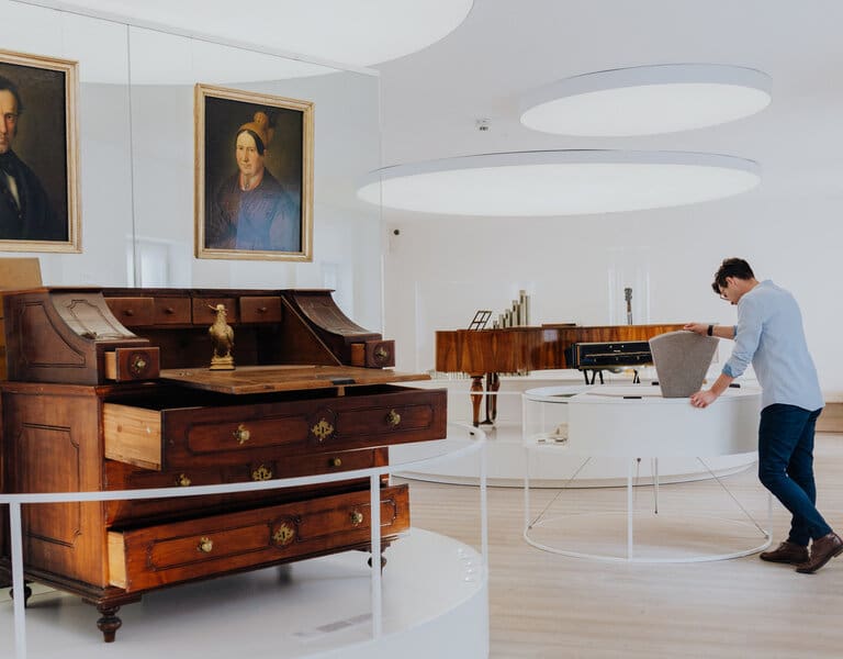 Im Vordergrund ist der Schreibtisch von Franz Xaver Gruber, sowie die Porträts von ihm und seiner Frau zu sehen. Im Hintergrund öffnet ein Besucher eine Schauklappe im Museum.