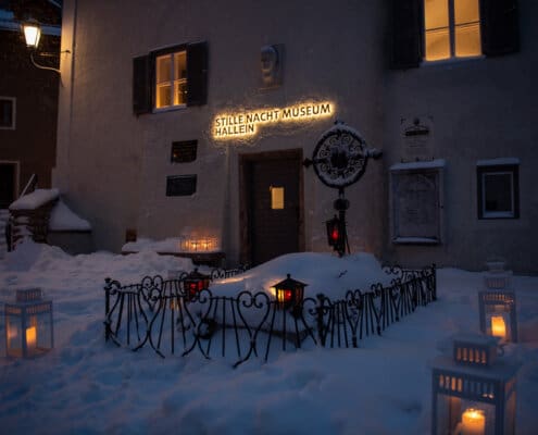 Das Grab von Franz Xaver Gruber unter einer Schneedecke, umgeben von Laternen, sowie der beleuchtete Eingang zum Stille Nacht Museum