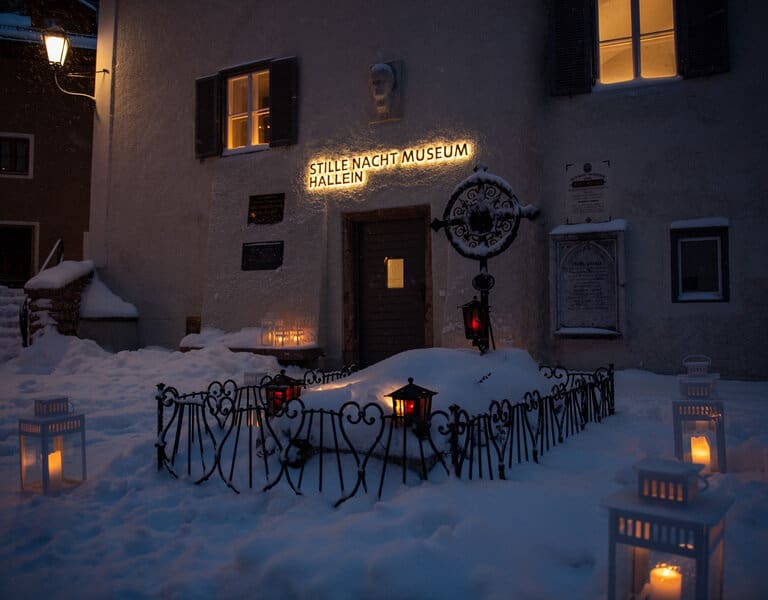 Das Grab von Franz Xaver Gruber unter einer Schneedecke, umgeben von Laternen, sowie der beleuchtete Eingang zum Stille Nacht Museum